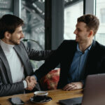 Two men engaged in a handshake at a table with a laptop, representing trust in business and real estate notarization.