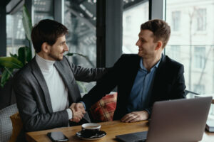 Two men engaged in a handshake at a table with a laptop, representing trust in business and real estate notarization.