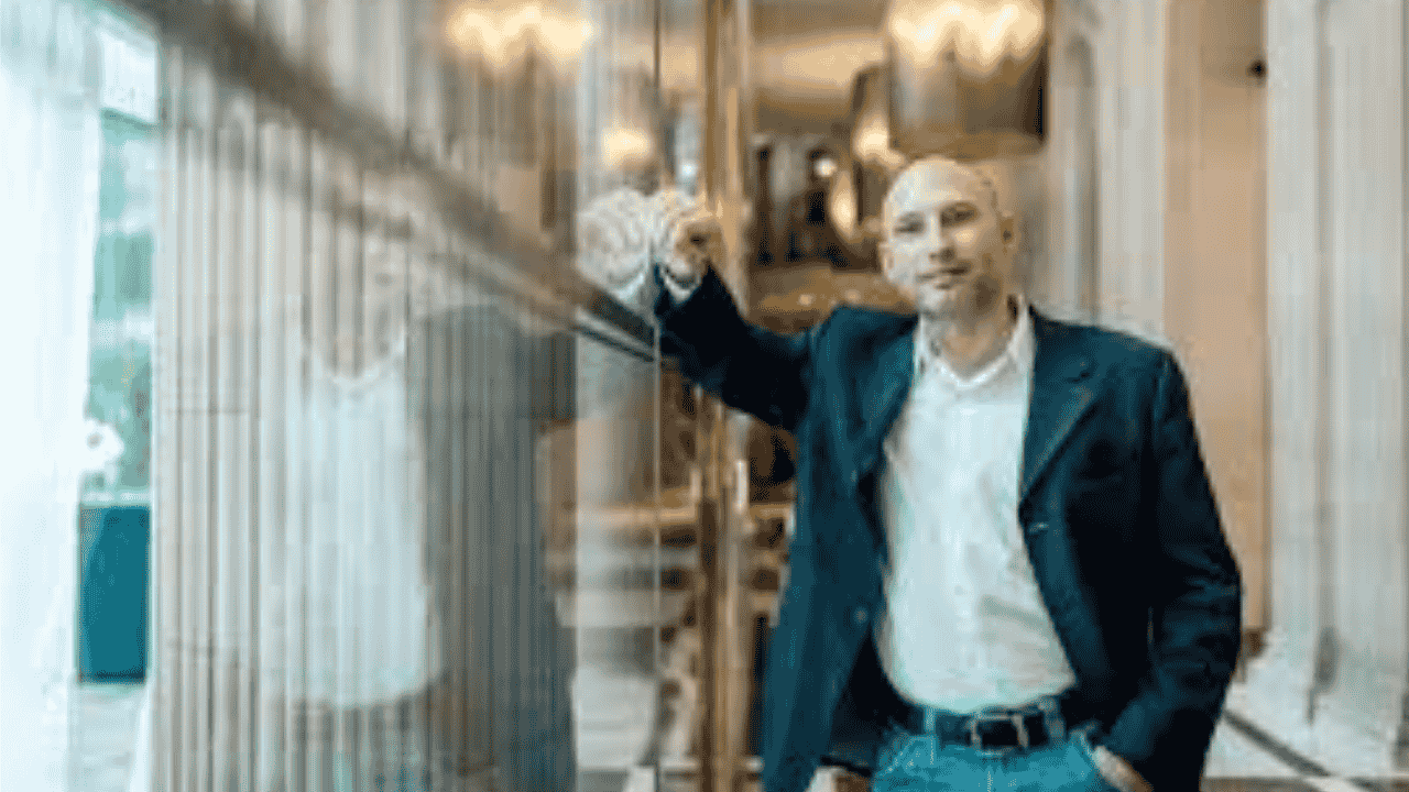 A man in a dark blazer and white shirt leaning against a glass railing in a grand, well-lit hallway with elegant architecture. He has a confident and approachable expression.