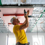 A skilled technician wearing safety gear, carefully installing a modern ceiling panel using advanced tools, in a well-lit interior space.