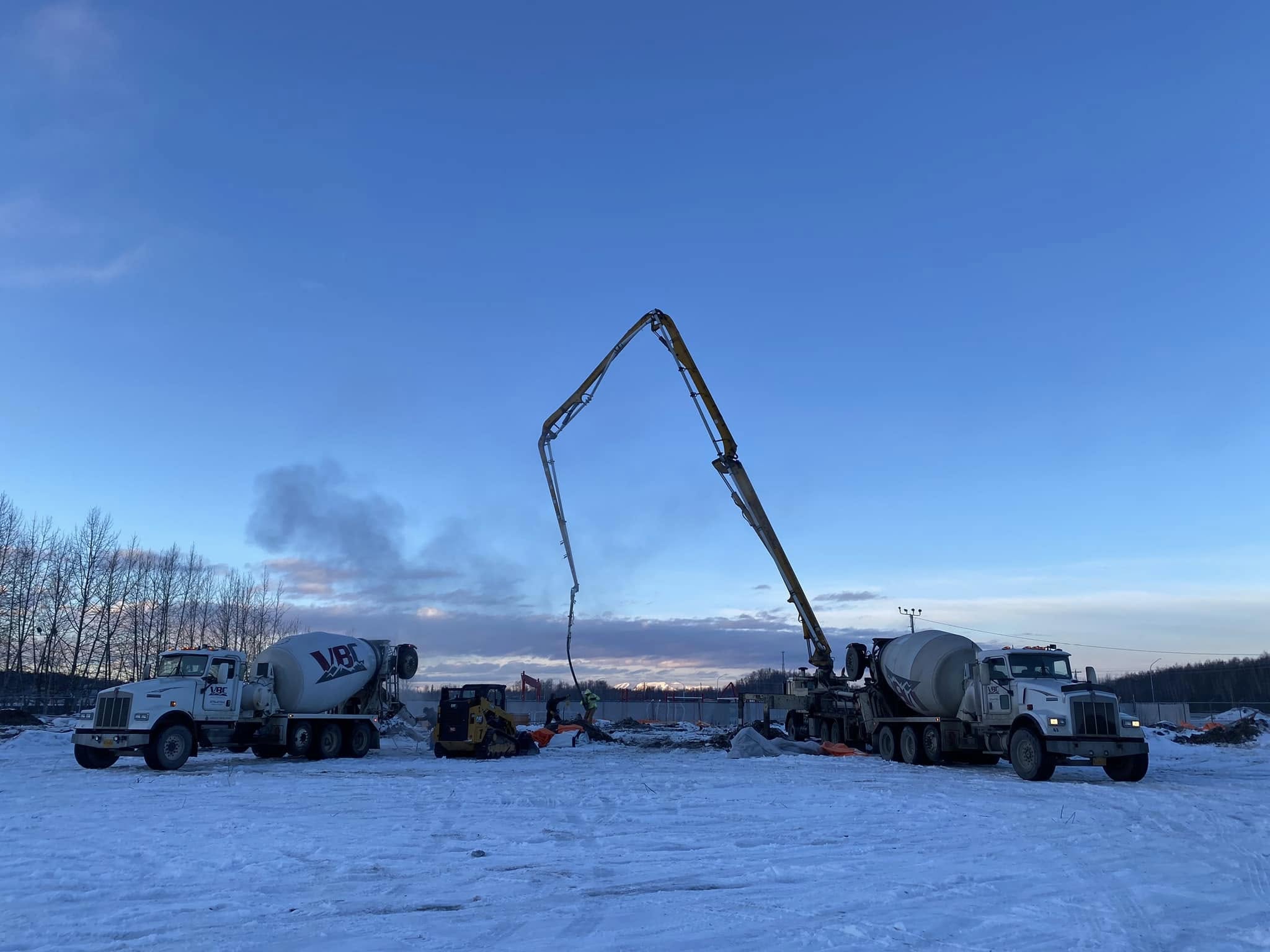 Concrete pumping at a snowy construction site with boom pump and mixer trucks working at dawn in Alaska.