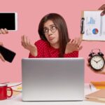 A woman sits at a desk with a laptop, looking overwhelmed, while several hands offer her a phone, tablet, reports, and a clock — symbolizing the stress of juggling multiple tasks and the need for productivity tools.